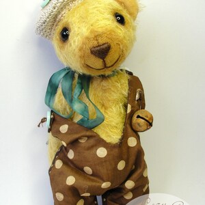 Puede incluir: Un oso de peluche hecho a mano que lleva un mono de lunares marrones y blancos, una cinta azul y un gorro de ganchillo. El oso tiene una campana atada a su mono y est&aacute; de pie sobre un fondo blanco. La imagen tambi&eacute;n incluye un logotipo para "Hasa's Cozy Corner Bears & More".