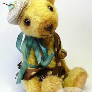 Puede incluir: Un oso de peluche hecho a mano que lleva un gorro de crochet con un bot&oacute;n azul. El oso est&aacute; sentado con las piernas cruzadas y lleva un vestido de lunares marrones y blancos con una cinta azul. El oso sostiene una peque&ntilde;a campana en su pata derecha. El oso est&aacute; sentado sobre una superficie blanca. La imagen incluye el texto "PDF Pattern of bear Louis" y "Lara's Cozy Corner Bears & more".