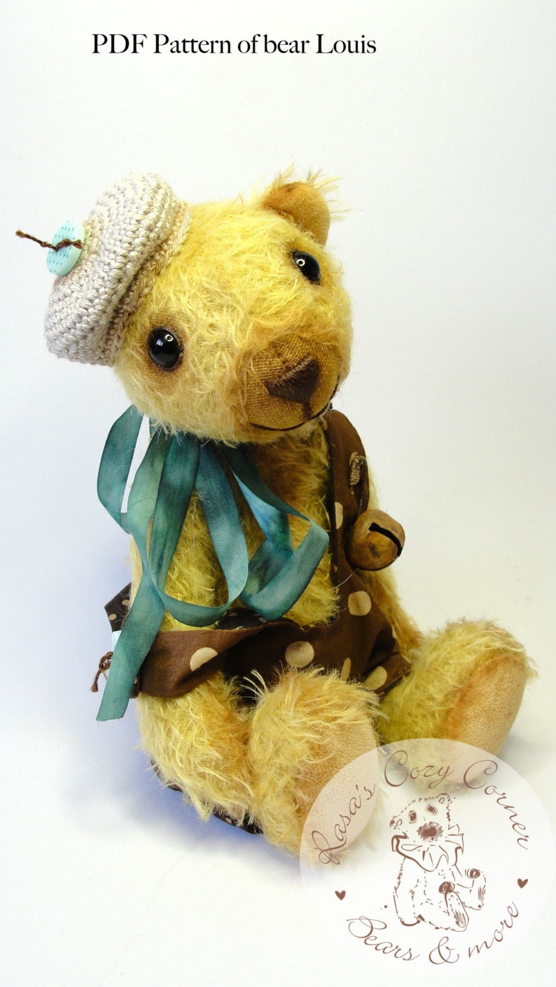 Puede incluir: Un oso de peluche hecho a mano que lleva un gorro de crochet con un bot&oacute;n azul. El oso est&aacute; sentado con las piernas cruzadas y lleva un vestido de lunares marrones y blancos con una cinta azul. El oso sostiene una peque&ntilde;a campana en su pata derecha. El oso est&aacute; sentado sobre una superficie blanca. La imagen incluye el texto "PDF Pattern of bear Louis" y "Lara's Cozy Corner Bears & more".