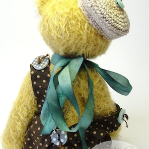 Puede incluir: Un oso de peluche amarillo que lleva un vestido de lunares marrones y blancos con una cinta verde, un sombrero de ganchillo y un bot&oacute;n azul claro. El oso est&aacute; sentado sobre una superficie blanca. La imagen incluye el texto "Hazel's Cozy Corner Bears & more".