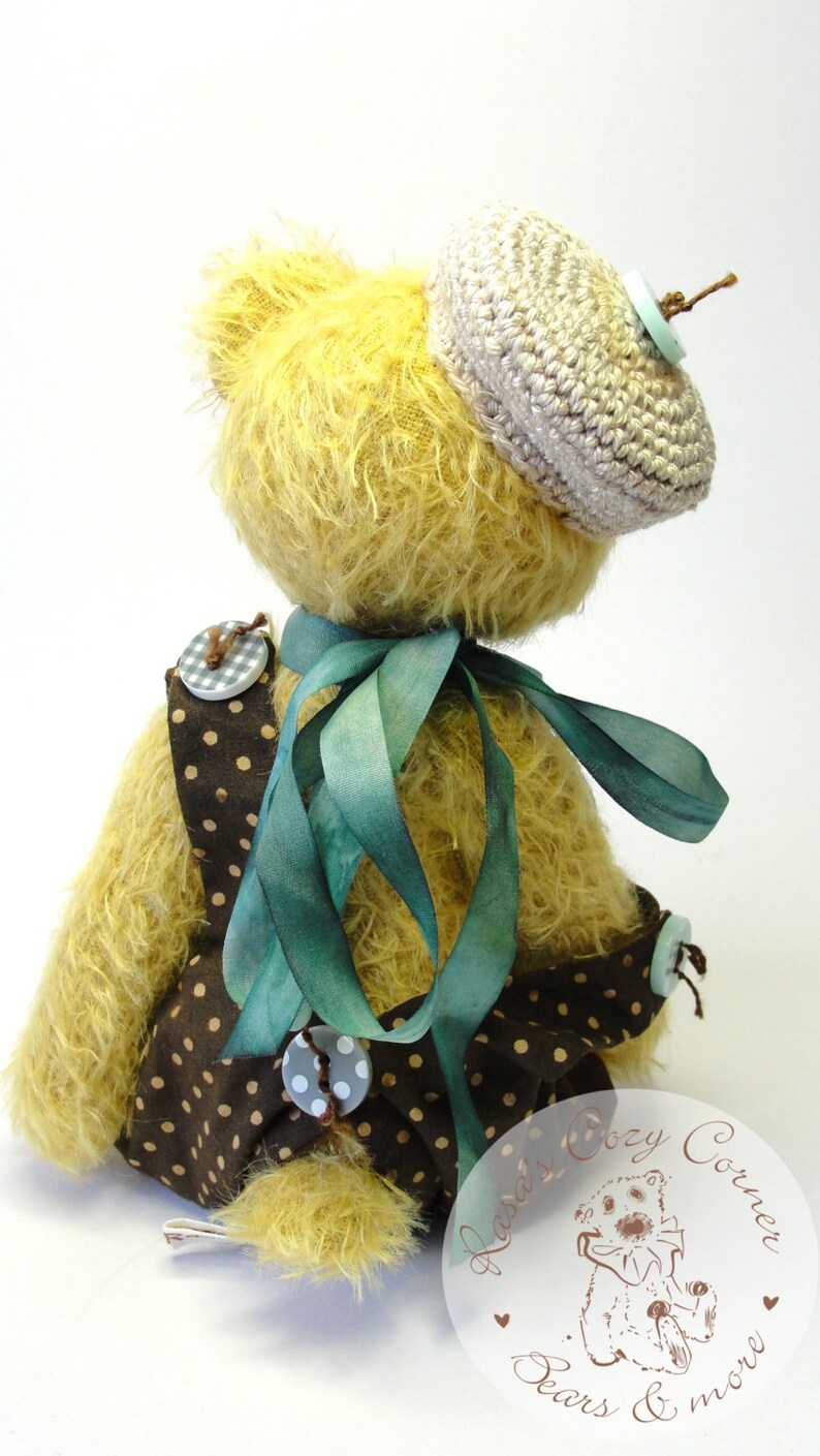 Puede incluir: Un oso de peluche amarillo que lleva un vestido de lunares marrones y blancos con una cinta verde, un sombrero de ganchillo y un bot&oacute;n azul claro. El oso est&aacute; sentado sobre una superficie blanca. La imagen incluye el texto "Hazel's Cozy Corner Bears & more".
