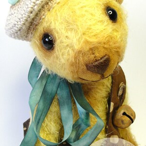 Puede incluir: Un oso de peluche amarillo que lleva un vestido de lunares marrones y blancos con una cinta turquesa, un sombrero de ganchillo y una campana. El oso est&aacute; sentado sobre un fondo blanco. El texto "Lasa's Cozy Corner Bears & more" es visible en la esquina inferior derecha de la imagen.