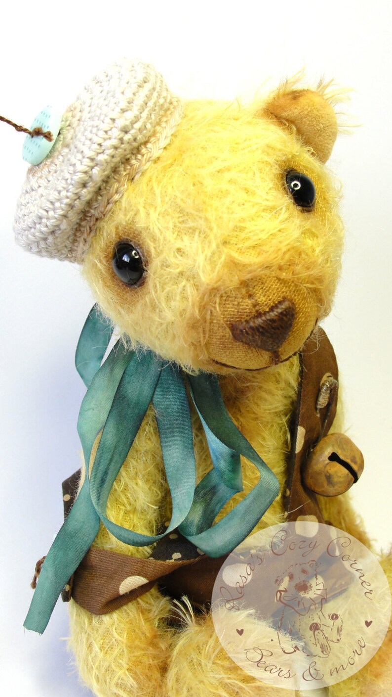 Puede incluir: Un oso de peluche amarillo que lleva un vestido de lunares marrones y blancos con una cinta turquesa, un sombrero de ganchillo y una campana. El oso est&aacute; sentado sobre un fondo blanco. El texto "Lasa's Cozy Corner Bears & more" es visible en la esquina inferior derecha de la imagen.