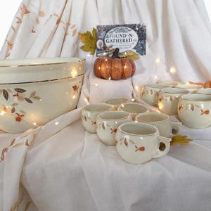 May include: A cream-colored ceramic bowl and matching cups with a fall-themed leaf design. The bowl has a gold rim, and the cups have handles. A small pumpkin and a sign that reads "FOUND-N-GATHERED CO" are also present.