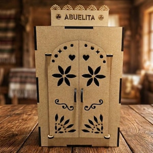 May include: A light brown wooden cabinet with two doors featuring laser-cut floral and heart patterns. The top section displays the text ABUELITA flanked by two small skull icons. The structure has a flat base and a decorative scalloped top edge.
