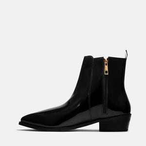 以下が含まれることがあります： ポインテッドトゥとローヒールが特徴の黒いアンクルブーツ。ブーツは、ゴールドカラーのプル付きのサイドジッパーを備えています。光沢のある仕上げが、ブーツに洗練されたモダンな外観を与えています。