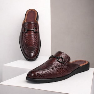 Könnte beinhalten: Ein Paar braune Leder-Slipper mit Reptilienhautstruktur. Jeder Schuh hat eine schwarze Schnalle und eine schwarze Sohle. Die Schuhe werden auf einer weißen Oberfläche vor einem neutralen Hintergrund präsentiert.