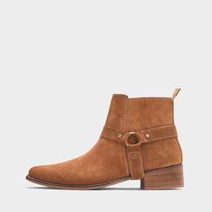 May include: A brown suede ankle boot with a pointed toe and a low stacked heel. The boot features a strap with a gold-colored metal ring and buckle detail around the ankle. The boot is approximately 6 inches tall.