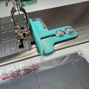 May include: A teal 3D-printed L-shaped sewing machine seam guide attached to the metal needle plate with two silver screws. The guide features a horizontal slot for adjustment and is positioned next to the presser foot and needle assembly.