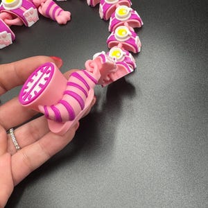 May include: A pink and purple articulated cat toy, featuring a striped body and a smiling face. The toy is made of plastic and has a whimsical design. The cat toy is approximately 7.6 cm long.