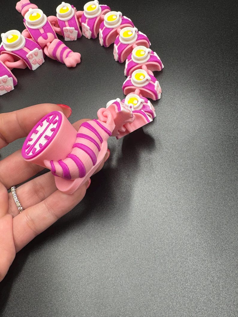 May include: A pink and purple articulated cat toy, featuring a striped body and a smiling face. The toy is made of plastic and has a whimsical design. The cat toy is approximately 7.6 cm long.