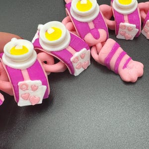 May include: A collection of pink and purple 3D-printed charms. Each charm features a white and yellow design, with a pink and purple base. Some charms have white panels with pink hearts.