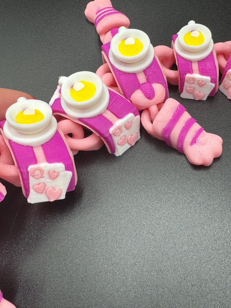 May include: A collection of pink and purple 3D-printed charms. Each charm features a white and yellow design, with a pink and purple base. Some charms have white panels with pink hearts.