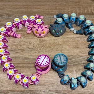 May include: A selection of articulated cat-themed toys in pink, blue, and grey. The toys feature cat heads, clock-faced designs, and segmented bodies, displayed on a wooden surface. The colour scheme is vibrant, with accents of yellow and white.