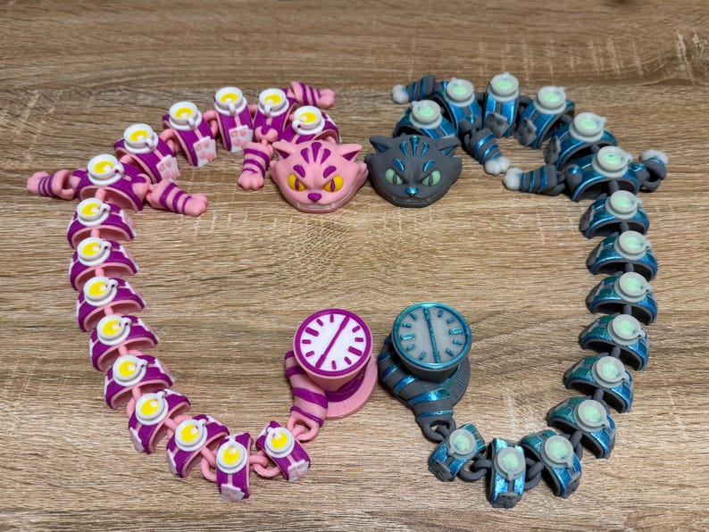 May include: A selection of articulated cat-themed toys in pink, blue, and grey. The toys feature cat heads, clock-faced designs, and segmented bodies, displayed on a wooden surface. The colour scheme is vibrant, with accents of yellow and white.