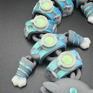 May include: A series of 3D-printed robot arm segments, each with a light blue and grey colour scheme. They have a circular, cup-like feature in the centre and white claw-like ends. The pieces are arranged on a dark surface, creating a chain-like appearance.