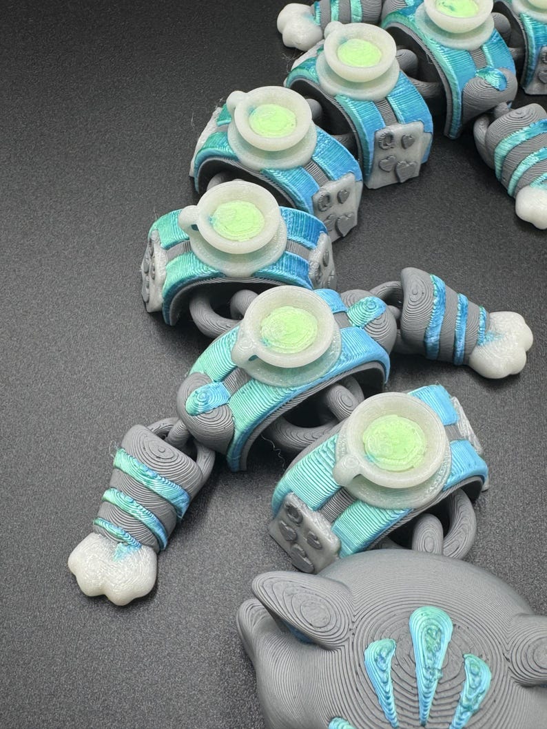May include: A series of 3D-printed robot arm segments, each with a light blue and grey colour scheme. They have a circular, cup-like feature in the centre and white claw-like ends. The pieces are arranged on a dark surface, creating a chain-like appearance.