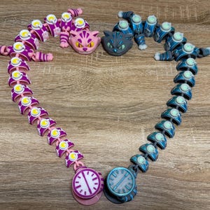 May include: Two articulated cat-themed toys in purple and blue, arranged in a heart shape. Each toy has a clock face at the bottom. The image also includes two cat heads, one pink and one grey.