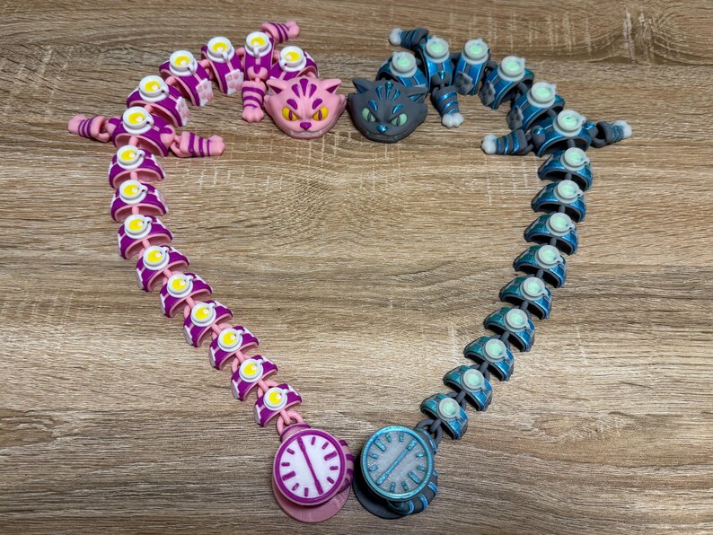 May include: Two articulated cat-themed toys in purple and blue, arranged in a heart shape. Each toy has a clock face at the bottom. The image also includes two cat heads, one pink and one grey.