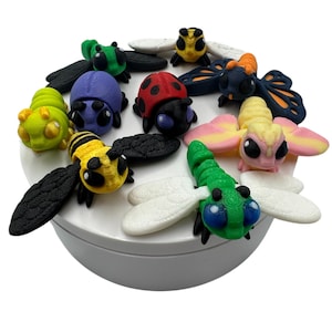 May include: A collection of colorful, 3D-printed insect figurines, including a ladybug, bee, butterfly, and dragonfly. The insects are in various colors, such as red, yellow, green, and blue, and are arranged on a white surface.