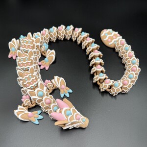 May include: A whimsical, articulated dragon figurine in shades of brown, white, pink, and blue. The dragon is made of connected segments, with a detailed head and tail. The design features decorative icing details, creating a playful and unique aesthetic.