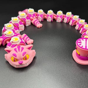 May include: A quirky, pink and purple Cheshire Cat-themed sculpture. The sculpture features a smiling cat head, a segmented body with teacups, and a teacup with a clock face. The design is inspired by Alice in Wonderland.