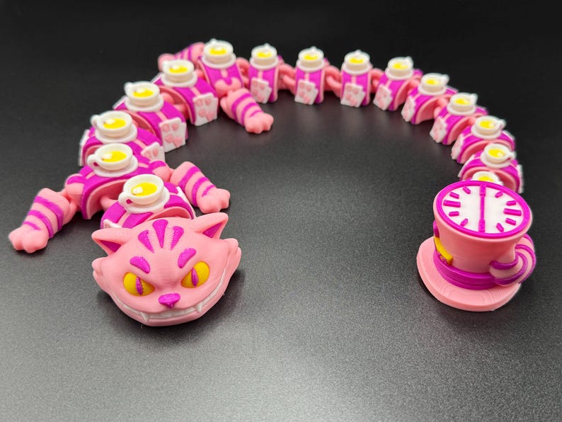 May include: A quirky, pink and purple Cheshire Cat-themed sculpture. The sculpture features a smiling cat head, a segmented body with teacups, and a teacup with a clock face. The design is inspired by Alice in Wonderland.