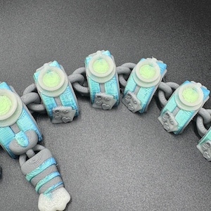 May include: A close-up of a chain featuring 3D-printed, abstract shapes in blue, teal, and grey. Each shape has a light green circular centre and is linked by a grey chain. The design is intricate, possibly a unique piece of jewellery or an accessory.