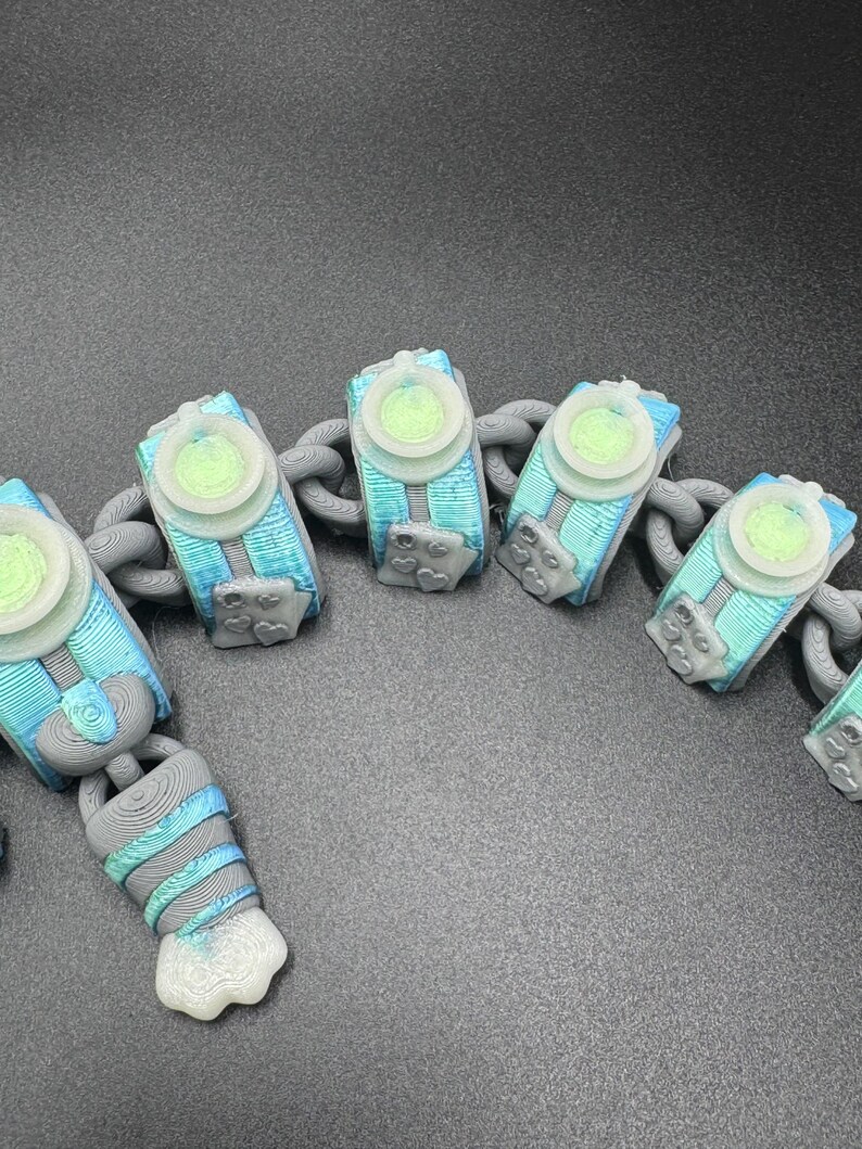 May include: A close-up of a chain featuring 3D-printed, abstract shapes in blue, teal, and grey. Each shape has a light green circular centre and is linked by a grey chain. The design is intricate, possibly a unique piece of jewellery or an accessory.