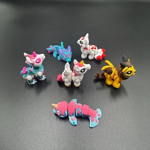 May include: A collection of colorful, handcrafted fantasy creatures. The figurines include a variety of mythical beings, such as unicorns, dragons, and other imaginative creatures. Each figure is uniquely designed with vibrant colors and intricate details, perfect for collectors or imaginative play.