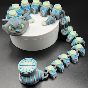 May include: An articulated cat toy with a Cheshire Cat head in grey with green eyes, and a segmented body in grey and blue. The toy includes a clock-like element. It is displayed on a white platform.