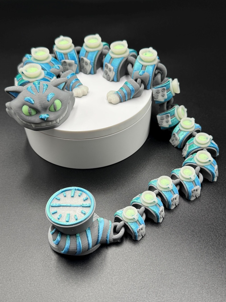 May include: An articulated cat toy with a Cheshire Cat head in grey with green eyes, and a segmented body in grey and blue. The toy includes a clock-like element. It is displayed on a white platform.