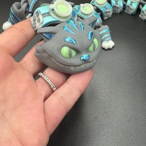 May include: A grey Cheshire Cat-themed articulated toy with blue and green details. The cat's face has a wide grin and green eyes. The toy is connected to other articulated pieces with teacups.