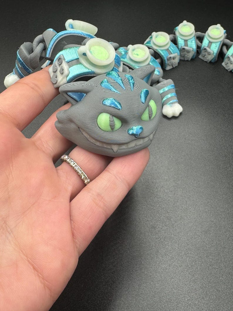 May include: A grey Cheshire Cat-themed articulated toy with blue and green details. The cat's face has a wide grin and green eyes. The toy is connected to other articulated pieces with teacups.