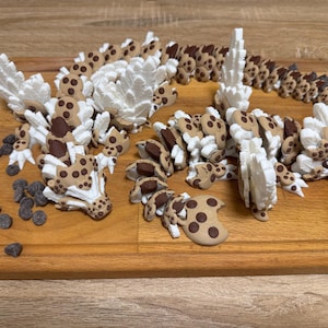 May include: A multi-part articulated dragon figurine constructed from tan and brown cookie-shaped segments with white, jagged, wing-like protrusions. The dragon features dark brown circular spots and is accompanied by several loose dark chocolate chips.