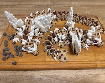 May include: A multi-part articulated dragon figurine constructed from tan and brown cookie-shaped segments with white, jagged, wing-like protrusions. The dragon features dark brown circular spots and is accompanied by several loose dark chocolate chips.