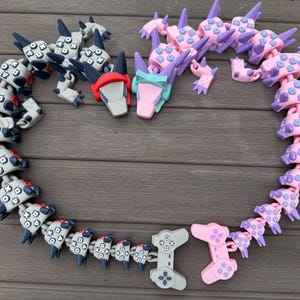May include: A collection of colorful, articulated toys arranged in a heart shape. The toys include gray and white controller-shaped pieces, and pink and purple dragon-like figures. The toys are made of plastic and are on a wooden surface.