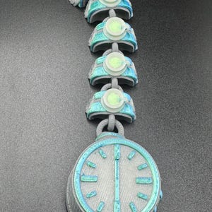 May include: A futuristic necklace with a large, round pendant resembling a pocket watch. The necklace is grey with teal and light green accents. The chain links have a similar design, creating a cohesive, sci-fi aesthetic.