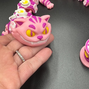 May include: A pink and purple articulated Cheshire Cat toy with yellow eyes and a wide grin. The toy is part of a series of linked pieces, including teacups and paws. The design is inspired by the Cheshire Cat from Alice in Wonderland.