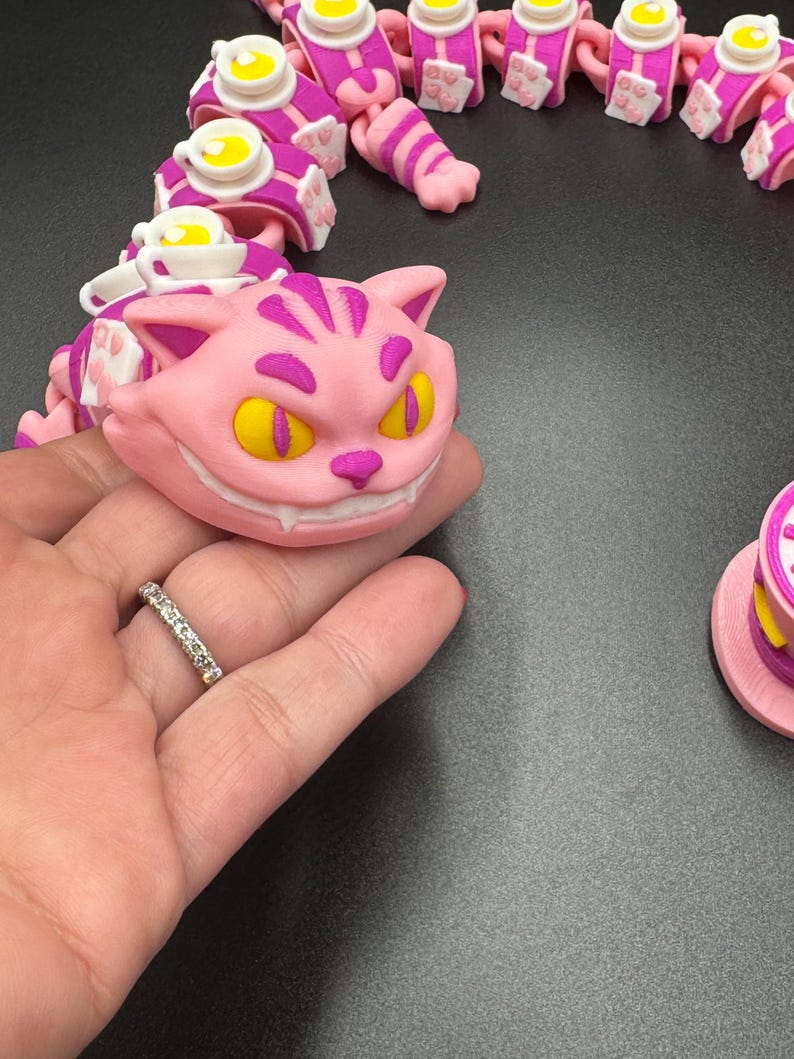 May include: A pink and purple articulated Cheshire Cat toy with yellow eyes and a wide grin. The toy is part of a series of linked pieces, including teacups and paws. The design is inspired by the Cheshire Cat from Alice in Wonderland.