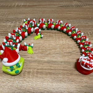 May include: A whimsical, articulated Christmas-themed dragon figurine. The dragon is primarily red, white, and green, with a Grinch-like head wearing a Santa hat. The body is segmented, resembling a chain of gifts.
