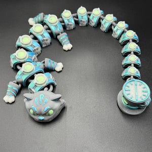 May include: A 3D-printed articulated cat toy in grey, blue, and green. The toy features a cat head, segmented body, and a clock-like tail end. The body segments have circular details and the cat's eyes glow green.