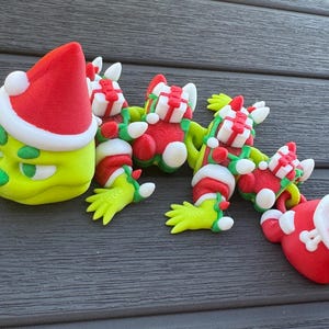 May include: A collection of whimsical Christmas-themed figurines. The figures are primarily green, red, and white, with a Grinch-like character wearing a Santa hat. The figurines are arranged on a dark gray surface.