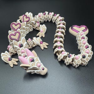 May include: A tan and white articulated dragon sculpture with pink and purple heart accents. The dragon has a heart-shaped tail and heart-shaped accents along its body. The dragon's head and feet are also visible.