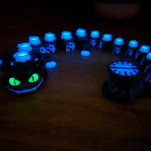 May include: A black, glowing caterpillar sculpture with blue and green highlights. The sculpture has a smiling cat face at one end and a circular design at the other. The caterpillar's body is segmented with glowing blue lights.