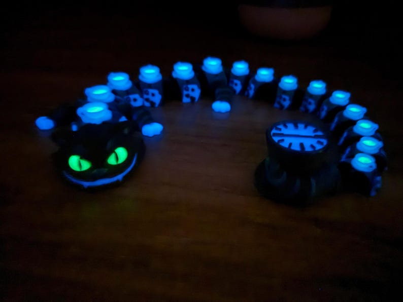 May include: A black, glowing caterpillar sculpture with blue and green highlights. The sculpture has a smiling cat face at one end and a circular design at the other. The caterpillar's body is segmented with glowing blue lights.