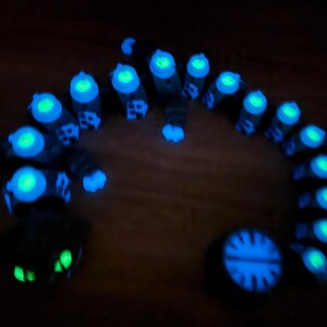 May include: A glowing, dark-coloured, segmented creature with bright blue and green accents. The creature is arranged in a curved shape, with a circular object and a face-like shape at either end. The image is taken in low light.