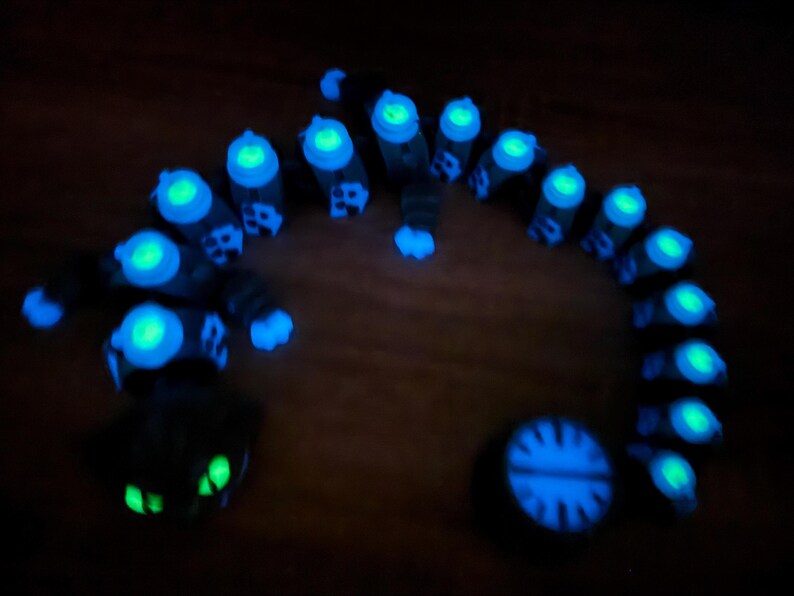 May include: A glowing, dark-coloured, segmented creature with bright blue and green accents. The creature is arranged in a curved shape, with a circular object and a face-like shape at either end. The image is taken in low light.