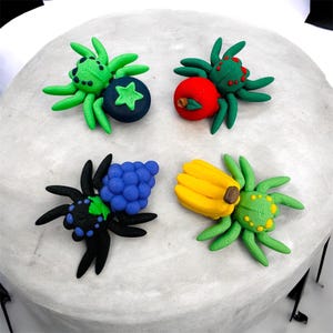 May include: Four colorful clay spider sculptures, each paired with a different fruit. The spiders are green, black, and red, with details like dots and textures. Fruits include a blueberry, apple, grapes, and bananas.