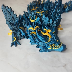 May include: A detailed, blue and gold dragon figurine. The dragon has intricate feather-like details and gold accents on its head, wings, and body. The figurine is posed on a white surface, showcasing its design.